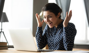 Excited young woman sitting at a desk using a laptop with a surprised joyful expression and upraised hands.