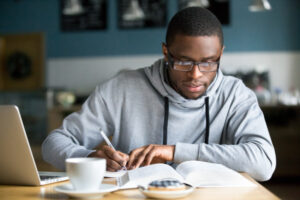 Focused male medical student studying at a coffee shop with an open book, laptop, coffee, and donut on table.