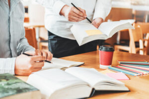 Open text books on a table with colored pencils lined up with two students, one seated and one standing, using pencils to point at the open books they are holding.