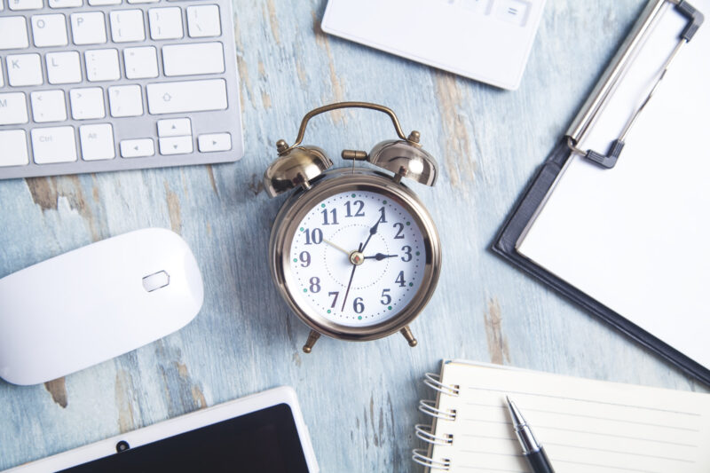 Silver clock surrounded by study items, notebook, clipboard, tablet, keyboard, mouse, and pen on a light blue background.