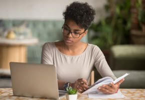 Young African american woman studying with a laptop on a desk while holding an open book.