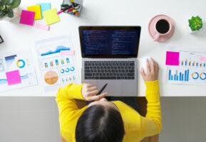 Overhead view of a woman in a yellow sweater seated at a desk with an open laptop, coffee, and colorful charts on papers on the desk.