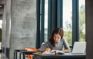 Asian student studies for the ABIM board exam by a large open window.