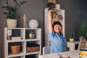 Female doctor in blue scrubs is stretching with a smile on her face while sitting at her desk in a bright and modern room, showing a moment of pain-relieving self care.