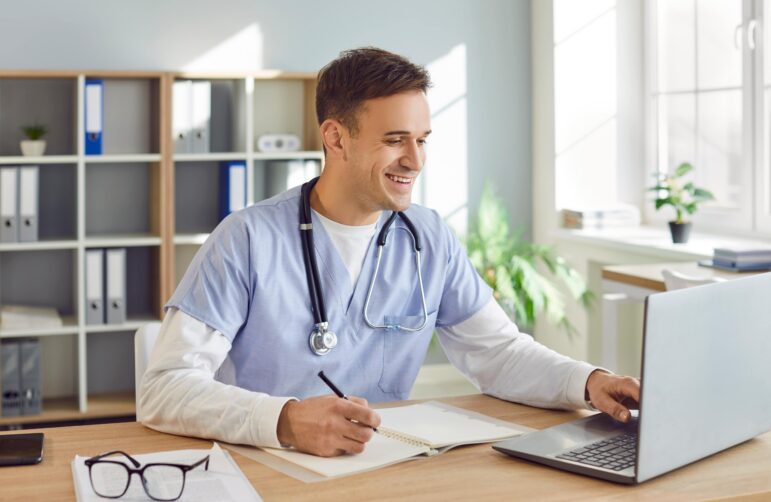 Physician studying for a medical board exam at a desk, reviewing notes and working on a laptop in a bright office setting.